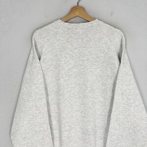 May include: A heather gray sweatshirt hangs on a wooden hanger. The sweatshirt has a crew neck and long sleeves. The fabric appears to be a soft, comfortable material. The garment is displayed against a plain gray background.