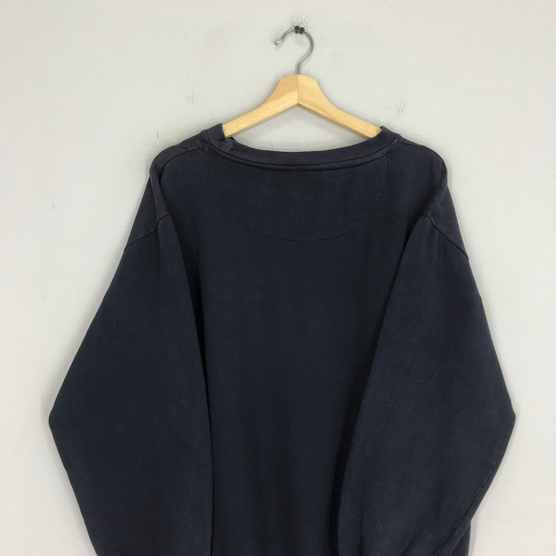 Puede incluir: Una sudadera azul oscuro con cuello redondo y mangas largas. La sudadera est&aacute; colgada de una percha de madera.