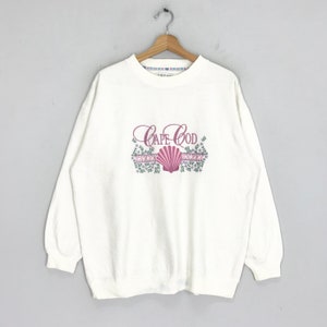 May include: White long-sleeve sweatshirt with a pink and green graphic of a seashell and the text "Cape Cod".