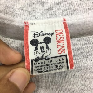 May include: Close-up of a gray sweatshirt tag with the Disney logo featuring Mickey Mouse. The tag reads "DESIGNS" and "MADE IN U.S.A." with fabric content and "ONE SIZE FITS ALL".