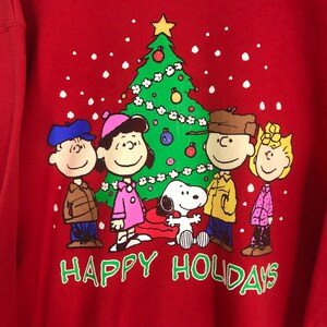 May include: Red sweatshirt with a Christmas tree, Snoopy, and the Peanuts gang. The text "Happy Holidays" is printed below the characters.