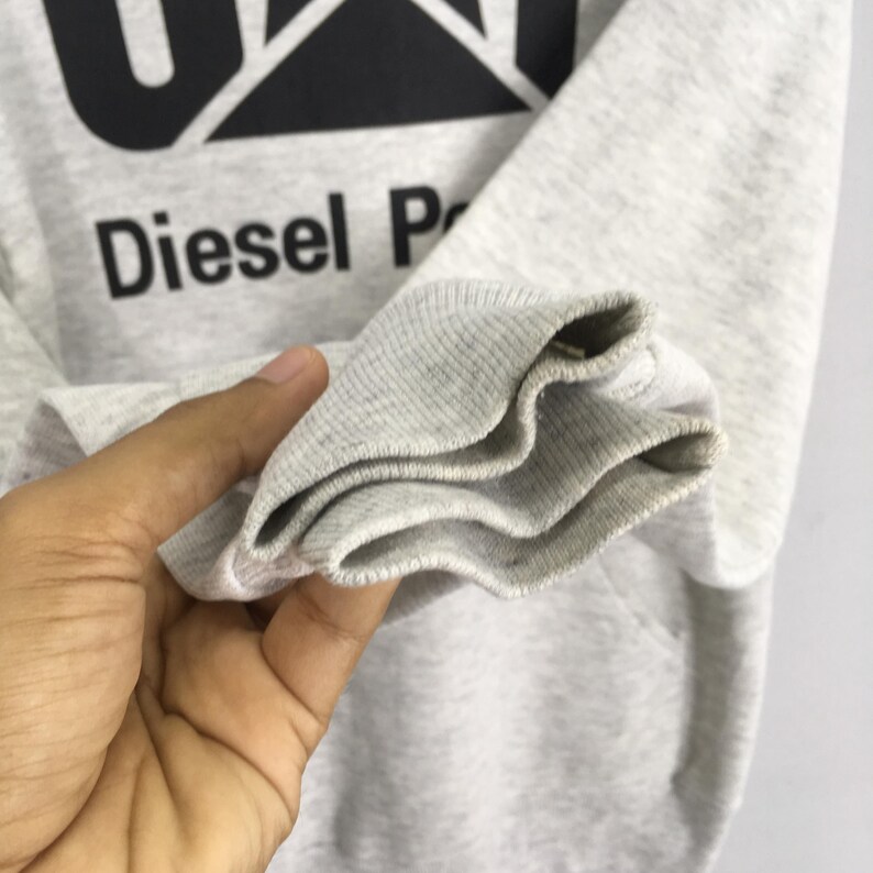 Puede incluir: Una sudadera de manga larga en color gris jaspeado claro con un estampado gr&aacute;fico negro en el pecho. El estampado muestra el texto CAT en una fuente negrita sobre las palabras Diesel Power. La prenda tiene pu&ntilde;os acanalados en las mangas.
