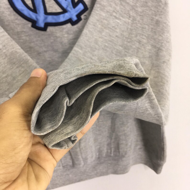 K&ouml;nnte beinhalten: Ein graues Sweatshirt mit einem hellblauen und marinefarbenen Emblem. Die &Auml;rmelmanschette ist umgeschlagen und zeigt die Innenseite des Stoffes. Das Sweatshirt besteht aus einem weichen, bequemen Material.