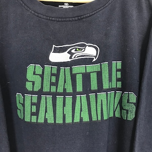 Puede incluir: Una camiseta negra de manga larga con un logotipo de los Seattle Seahawks en verde y blanco. El logotipo representa un p&aacute;jaro estilizado con un pico y ojos verdes. El texto "SEATTLE SEAHAWKS" est&aacute; impreso en verde debajo del logotipo.