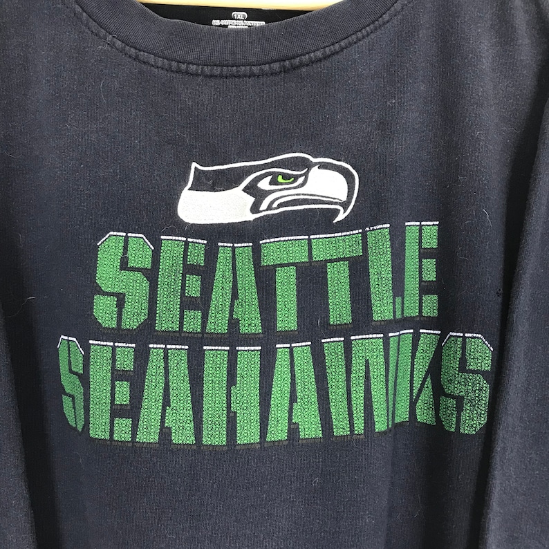 Puede incluir: Una camiseta negra de manga larga con un logotipo de los Seattle Seahawks en verde y blanco. El logotipo representa un p&aacute;jaro estilizado con un pico y ojos verdes. El texto "SEATTLE SEAHAWKS" est&aacute; impreso en verde debajo del logotipo.