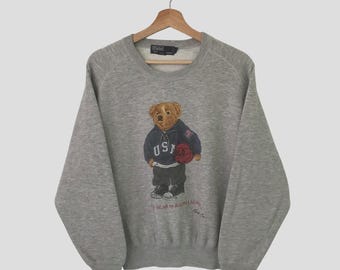 May include: Gray sweatshirt with a graphic of a bear wearing a blue and red USA shirt and holding a basketball. The bear is wearing black pants and sneakers. The text "Polo Bear by Ralph Lauren" is printed below the bear.
