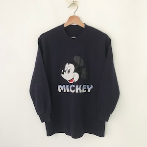 Peut inclure: Un sweat-shirt à manches longues bleu marine avec une grande image brodée du visage de Mickey Mouse et le mot "MICKEY" dans une police de caractères bleu et blanc dégradée.