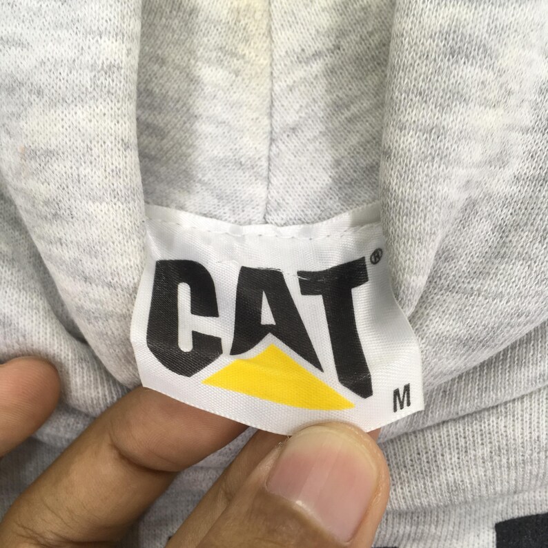 Puede incluir: Una sudadera con capucha de tela jaspeada gris claro que presenta una etiqueta rectangular blanca en el cuello. La etiqueta muestra el texto negro CAT con un tri&aacute;ngulo amarillo debajo y la letra M impresa en negro a la derecha.