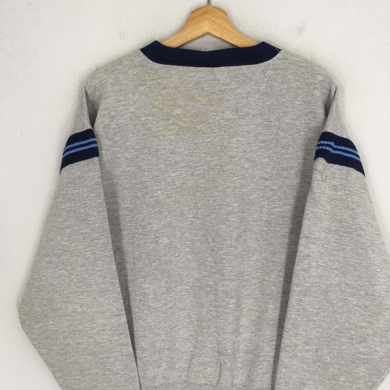 K&ouml;nnte beinhalten: Ein graues Sweatshirt mit marineblauem Kragen und blauen Streifen auf den &Auml;rmeln. Das Sweatshirt h&auml;ngt an einem Holzb&uuml;gel vor einem wei&szlig;en Hintergrund. Die &Auml;rmel haben ein dunkelblaues Band mit zwei hellblauen Streifen.