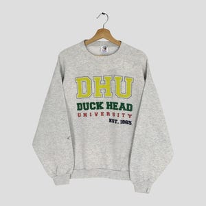 May include: A light gray sweatshirt with the text "DHU DUCK HEAD UNIVERSITY EST. 1865" in yellow, green, and red. The sweatshirt is hanging on a wooden hanger.