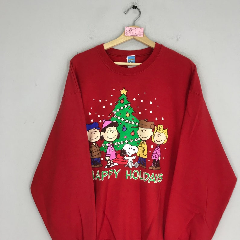 May include: Red sweatshirt with a Christmas tree and the Peanuts characters, including Snoopy, Charlie Brown, Lucy, Linus, and Sally. The text "Happy Holidays" is printed below the characters.