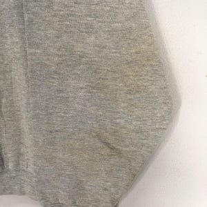 K&ouml;nnte beinhalten: Nahaufnahme eines melierten grauen Sweatshirts. Der Stoff hat ein strukturiertes Aussehen mit subtilen Farbvariationen. Das Kleidungsst&uuml;ck weist Gebrauchsspuren auf, mit sichtbaren Verf&auml;rbungen auf dem Stoff.