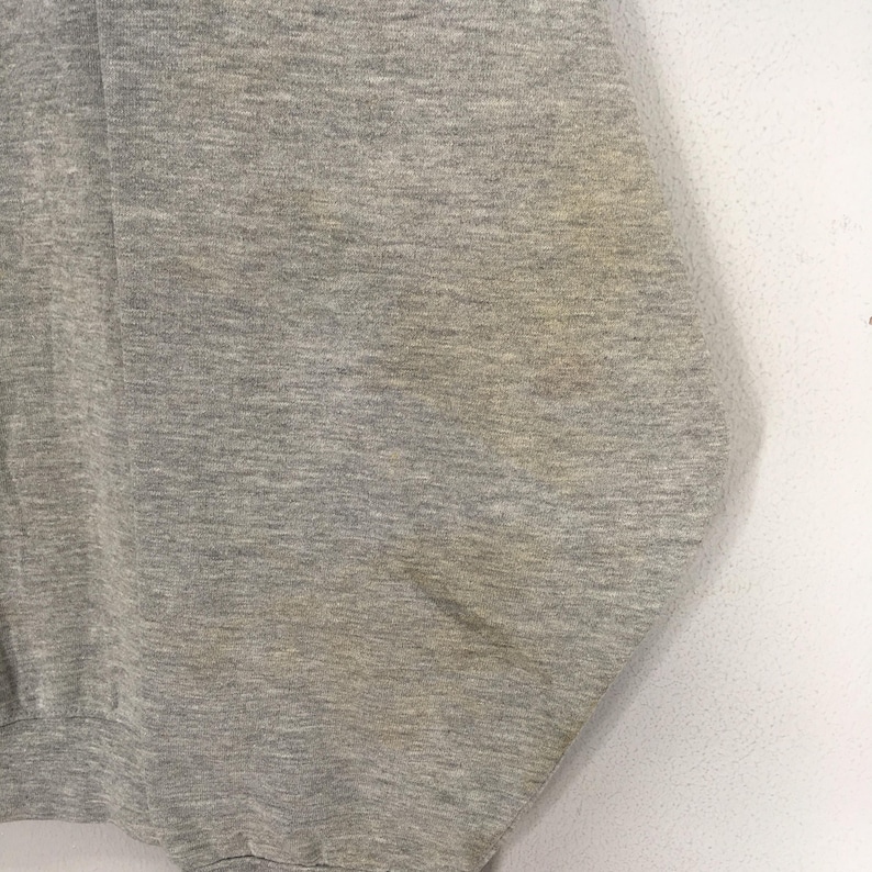 K&ouml;nnte beinhalten: Nahaufnahme eines melierten grauen Sweatshirts. Der Stoff hat ein strukturiertes Aussehen mit subtilen Farbvariationen. Das Kleidungsst&uuml;ck weist Gebrauchsspuren auf, mit sichtbaren Verf&auml;rbungen auf dem Stoff.