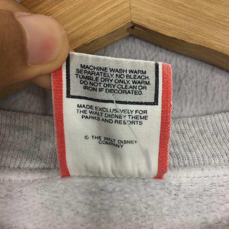 May include: A close-up of a tag from a gray sweatshirt. The tag is white with red trim and features care instructions and the text "MADE EXCLUSIVELY FOR THE WALT DISNEY THEME PARKS AND RESORTS" and "THE WALT DISNEY COMPANY".