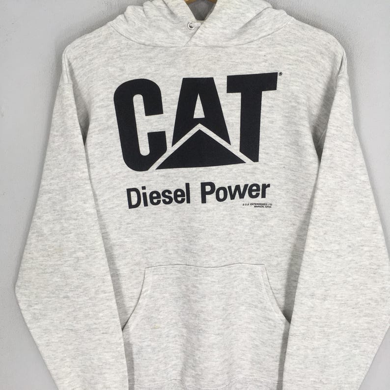 Puede incluir: Una sudadera con capucha de manga larga en color gris jaspeado claro que presenta un gran logotipo CAT negro impreso en el pecho. Debajo del logotipo, el texto dice Diesel Power en letras negras, con un peque&ntilde;o texto de copyright bajo la palabra Power.