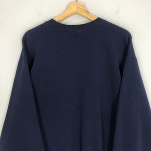 May include: A navy blue sweatshirt hanging on a wooden hanger. The sweatshirt has a crew neck and long sleeves. The fabric appears to be a soft, medium-weight material. The background is a plain, off-white wall.