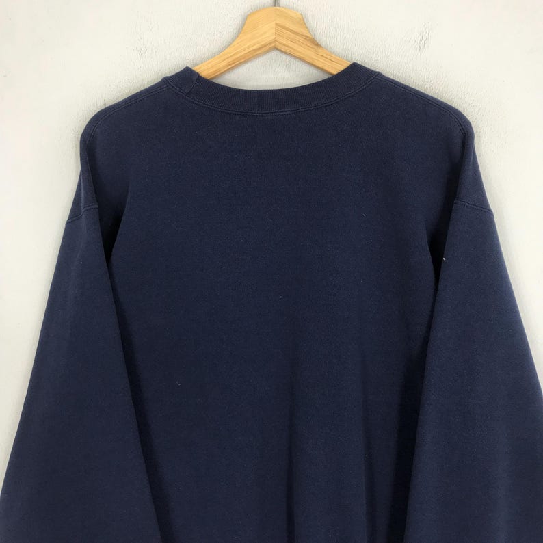 May include: A navy blue sweatshirt hanging on a wooden hanger. The sweatshirt has a crew neck and long sleeves. The fabric appears to be a soft, medium-weight material. The background is a plain, off-white wall.