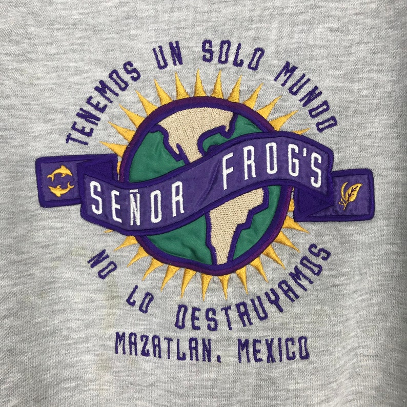 May include: A gray sweatshirt with a purple and yellow embroidered logo. The logo features a globe with a purple banner that reads "Se&ntilde;or Frog's". The text around the globe reads "Tenemos un solo mundo no lo destruya mos Mazatlan, Mexico".