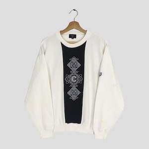 Puede incluir: Sudadera de color crema con un panel vertical negro en el centro. El panel presenta un diseño bordado con las letras "MCM" en un patrón de diamantes. Las mangas son largas y la sudadera está colgada de una percha de madera.