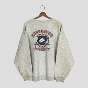 May include: A light grey sweatshirt featuring the text "RODBUSTER SCHOOL OF MAR MAC UNIVERSITY 2006" in a circular design. The design includes a blue, black, and white emblem. The sweatshirt is suspended from a wooden hanger.