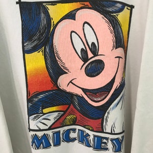May include: A white t-shirt with a colorful cartoon image of Mickey Mouse. The image is framed in a yellow, orange, and red border. The text "MICKEY" is printed in blue below the image.