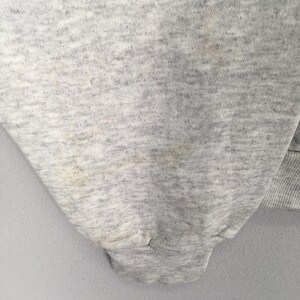 May include: Close-up of a light gray sweatshirt sleeve. The fabric has a textured appearance with some discoloration and stains. The cuff is ribbed and the overall garment appears to be well-worn.