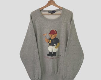 May include: A gray sweatshirt with a large graphic of a bear wearing a red helmet and holding a polo mallet. The bear is wearing a blue and yellow polo shirt with the number 2 on it. The text "POLO RALPH LAUREN" is printed below the bear.