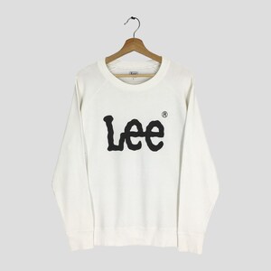 Könnte beinhalten: Ein weißes Sweatshirt mit Rundhalsausschnitt und dem schwarzen Wort "Lee" auf der Vorderseite. Das Sweatshirt hängt an einem Holzbügel. Das "Lee"-Logo ist in einer fetten, abgerundeten Schriftart.