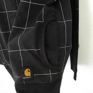 May include: Black zip-up hoodie with a white grid pattern and a Carhartt logo patch. The hoodie has a kangaroo pocket and a ribbed hem.