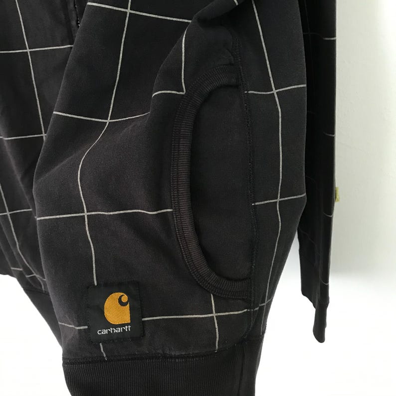 May include: Black zip-up hoodie with a white grid pattern and a Carhartt logo patch. The hoodie has a kangaroo pocket and a ribbed hem.