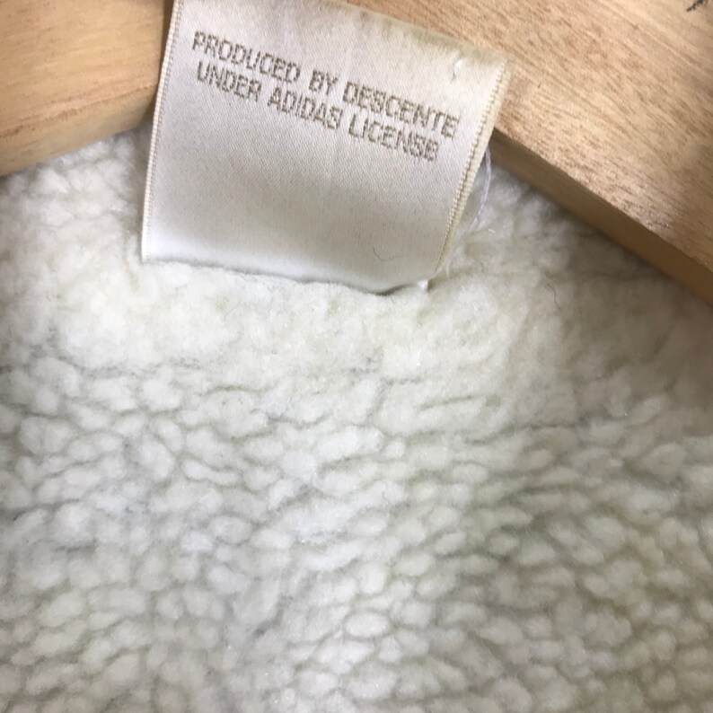 May include: White tag with the text "PRODUCED BY DESCENTE UNDER ADIDAS LICENSE" on a white fleece fabric.