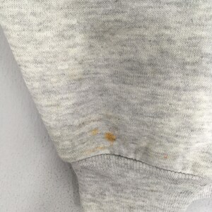 Puede incluir: Una manga de tela jaspeada gris claro con un pu&ntilde;o acanalado. El material muestra una mancha visible de color amarillo parduzco ubicada cerca del &aacute;rea del pu&ntilde;o, mostrando la textura del tejido de punto y la ubicaci&oacute;n espec&iacute;fica de la mancha.