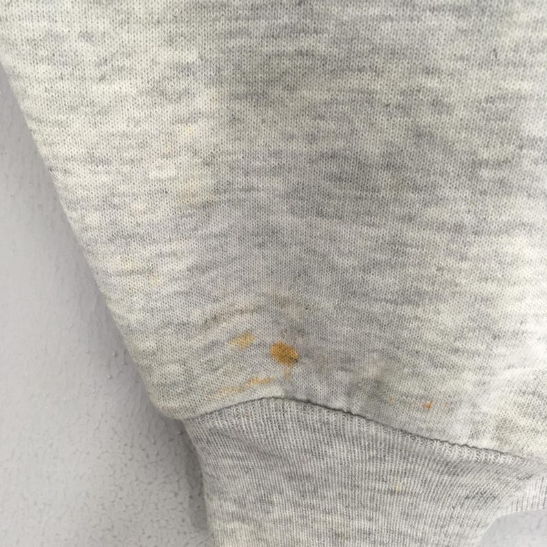 Puede incluir: Una manga de tela jaspeada gris claro con un pu&ntilde;o acanalado. El material muestra una mancha visible de color amarillo parduzco ubicada cerca del &aacute;rea del pu&ntilde;o, mostrando la textura del tejido de punto y la ubicaci&oacute;n espec&iacute;fica de la mancha.