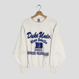 Może przedstawiać: Jasnoszara bluza z napisem "Duke Univ. Blue Devils" w niebieskim kolorze. Poniżej niebieskie logo i napis "DUKE NORTH CAROLINA". Bluza wisi na drewnianym wieszaku.