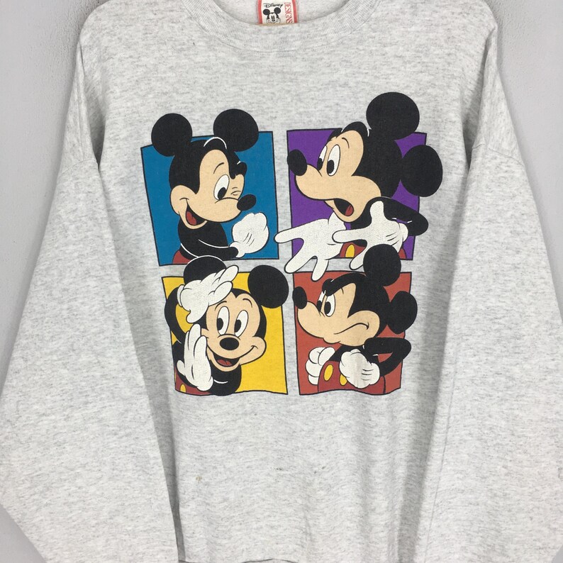 May include: A light gray sweatshirt featuring a colorful graphic of Mickey Mouse in four different poses. The graphic is divided into four squares with blue, purple, yellow, and red backgrounds. The sweatshirt has long sleeves and a crew neck.