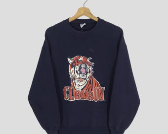 May include: Navy blue sweatshirt featuring a roaring tiger graphic in orange, white, and black, with the word "CLEMSON" in orange block letters. The sweatshirt has a crew neck and long sleeves, suitable for casual wear.
