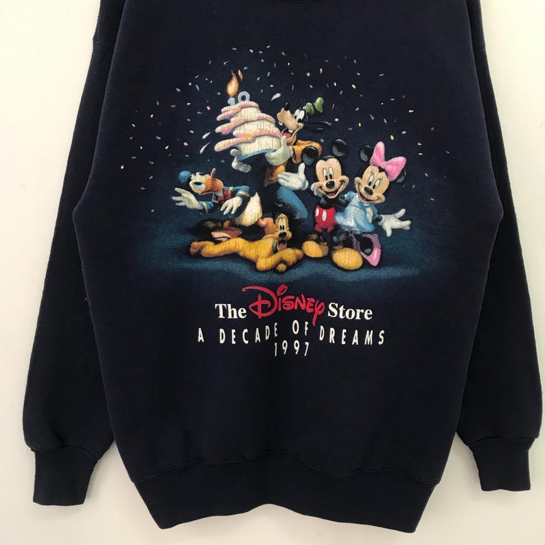 May include: A navy blue sweatshirt with a colorful graphic of Mickey Mouse and friends celebrating a birthday. The text "The Disney Store A Decade of Dreams 1997" is printed below the graphic.