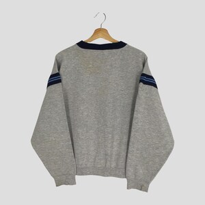 K&ouml;nnte beinhalten: Ein graues Sweatshirt mit marineblauem Kragen und zwei marineblauen Streifen auf jedem &Auml;rmel. Das Sweatshirt h&auml;ngt an einem Holzb&uuml;gel vor einem hellen Hintergrund. Die &Auml;rmel sind lang und das Material scheint ein weicher Stoff zu sein.