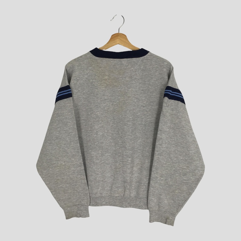 K&ouml;nnte beinhalten: Ein graues Sweatshirt mit marineblauem Kragen und zwei marineblauen Streifen auf jedem &Auml;rmel. Das Sweatshirt h&auml;ngt an einem Holzb&uuml;gel vor einem hellen Hintergrund. Die &Auml;rmel sind lang und das Material scheint ein weicher Stoff zu sein.