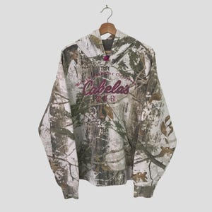 Pode incluir: Uma jaqueta de moletom com capuz camuflada branca, verde e marrom com um logotipo "Cabelas" rosa e o texto "World's Foremost Outfitter" na frente.
