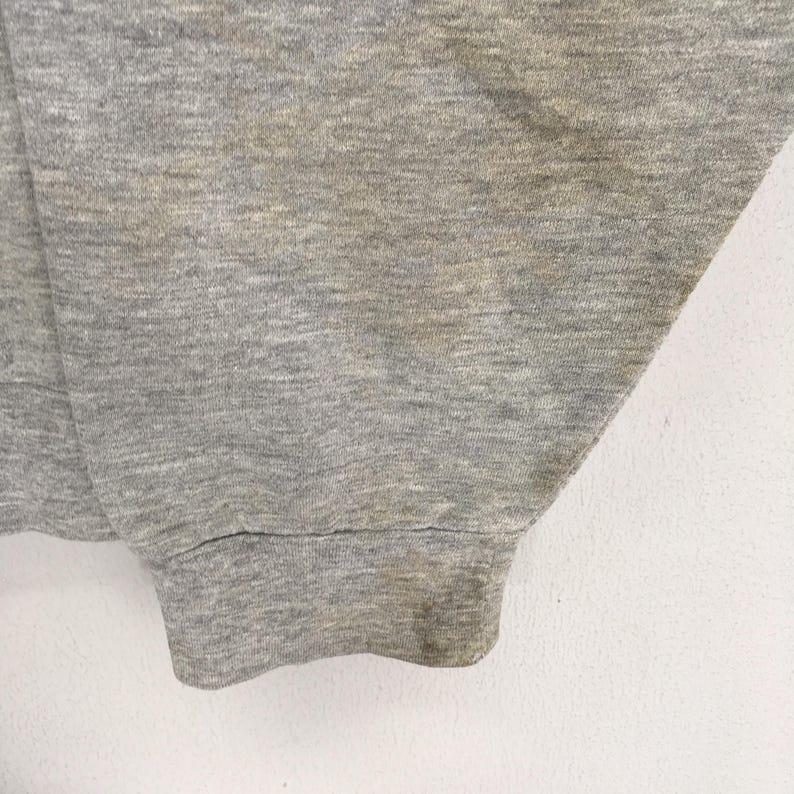 K&ouml;nnte beinhalten: Nahaufnahme eines grauen Sweatshirt-&Auml;rmels. Der Stoff hat eine melierte Textur mit einigen Verf&auml;rbungen. Das B&uuml;ndchen ist gerippt und weist Abnutzungserscheinungen auf. Der Hintergrund ist eine schlichte wei&szlig;e Oberfl&auml;che.