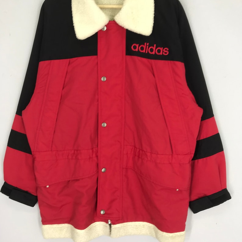 May include: A red and black vintage Adidas jacket with a white faux fur collar and a black and red striped sleeve. The jacket has a zipper closure and button accents. The Adidas logo is embroidered in red on the chest.