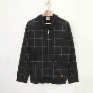 May include: Black hooded sweatshirt with a white grid pattern and a zipper closure. The sweatshirt has a small Carhartt logo patch on the bottom right side.