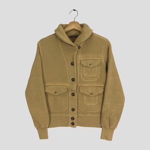 Puede incluir: Chaqueta color canela con cuello ancho, cuatro bolsillos delanteros y cierre de botones. La chaqueta tiene mangas largas con puños acanalados y un aspecto ligeramente descolorido. Cuelga de una percha de madera sobre un fondo gris claro.