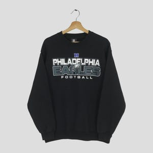 Könnte beinhalten: Schwarzes Sweatshirt mit dem Text "PHILADELPHIA EAGLES FOOTBALL" in Weiß, Grau und Blau. Das Sweatshirt hängt an einem Holzkleiderbügel.
