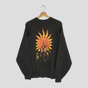 May include: Black sweatshirt with a vibrant sun graphic on the back. The sun design features yellow and orange hues, with a stylized tree and text below. The sweatshirt is displayed on a wooden hanger.