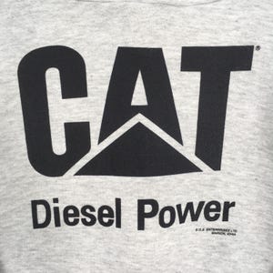 Puede incluir: Una prenda de tela gris claro jaspeada con un gran logotipo CAT negro impreso en el centro. Debajo del logotipo, el texto dice Diesel Power en negro, con un texto m&aacute;s peque&ntilde;o que indica S.O.S. ENTERPRISES LTD. MARION, IOWA abajo a la derecha.
