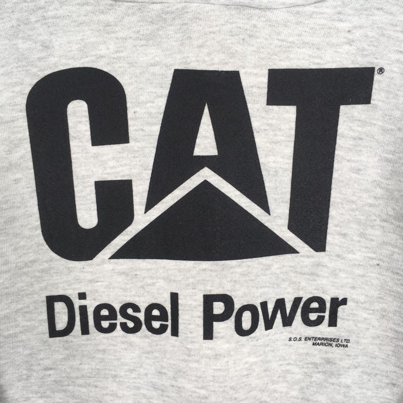 Puede incluir: Una prenda de tela gris claro jaspeada con un gran logotipo CAT negro impreso en el centro. Debajo del logotipo, el texto dice Diesel Power en negro, con un texto m&aacute;s peque&ntilde;o que indica S.O.S. ENTERPRISES LTD. MARION, IOWA abajo a la derecha.