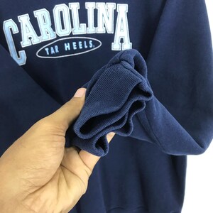 May include: Navy blue sweatshirt with the word "CAROLINA" in white and light blue lettering. Below the word "CAROLINA" is the text "TAR HEELS" in white lettering inside of an oval. The sleeve cuff is visible.