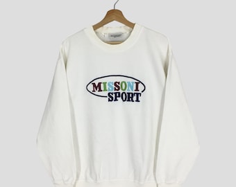 Puede incluir: Una sudadera blanca de cuello redondo con el texto "MISSONI SPORT" bordado en un diseño ovalado azul, verde y marrón. La sudadera está colgada de una percha de madera.
